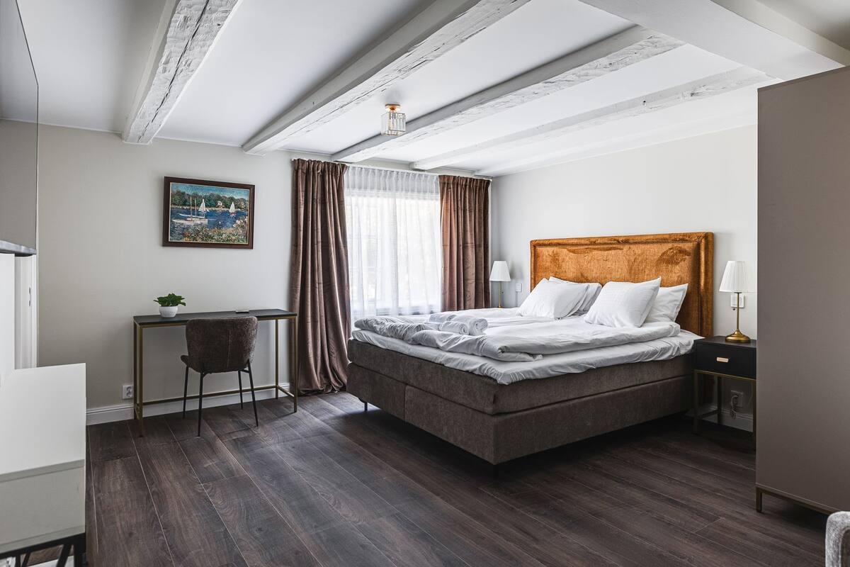 Un dormitorio con cama, escritorio, silla, lámpara y un cuadro en la pared.