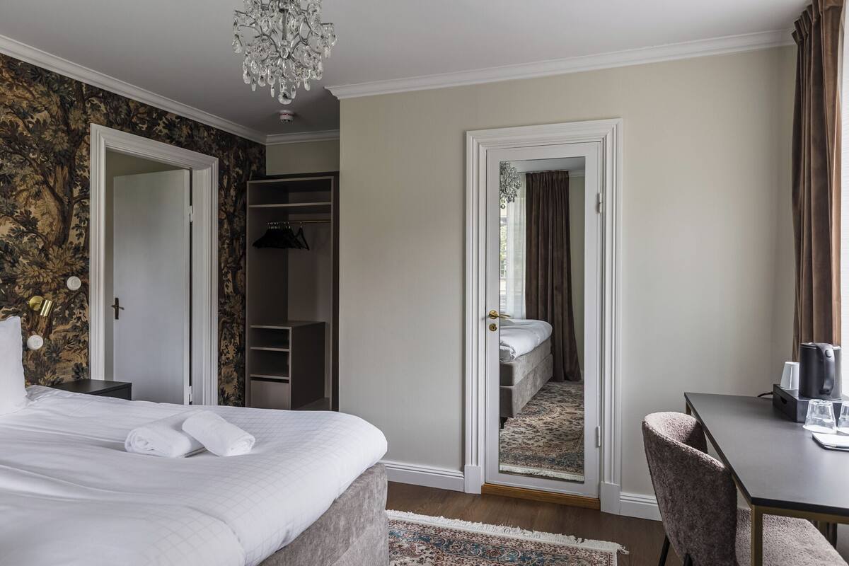 Una habitación de hotel con cama, escritorio, silla, armario y espejo.