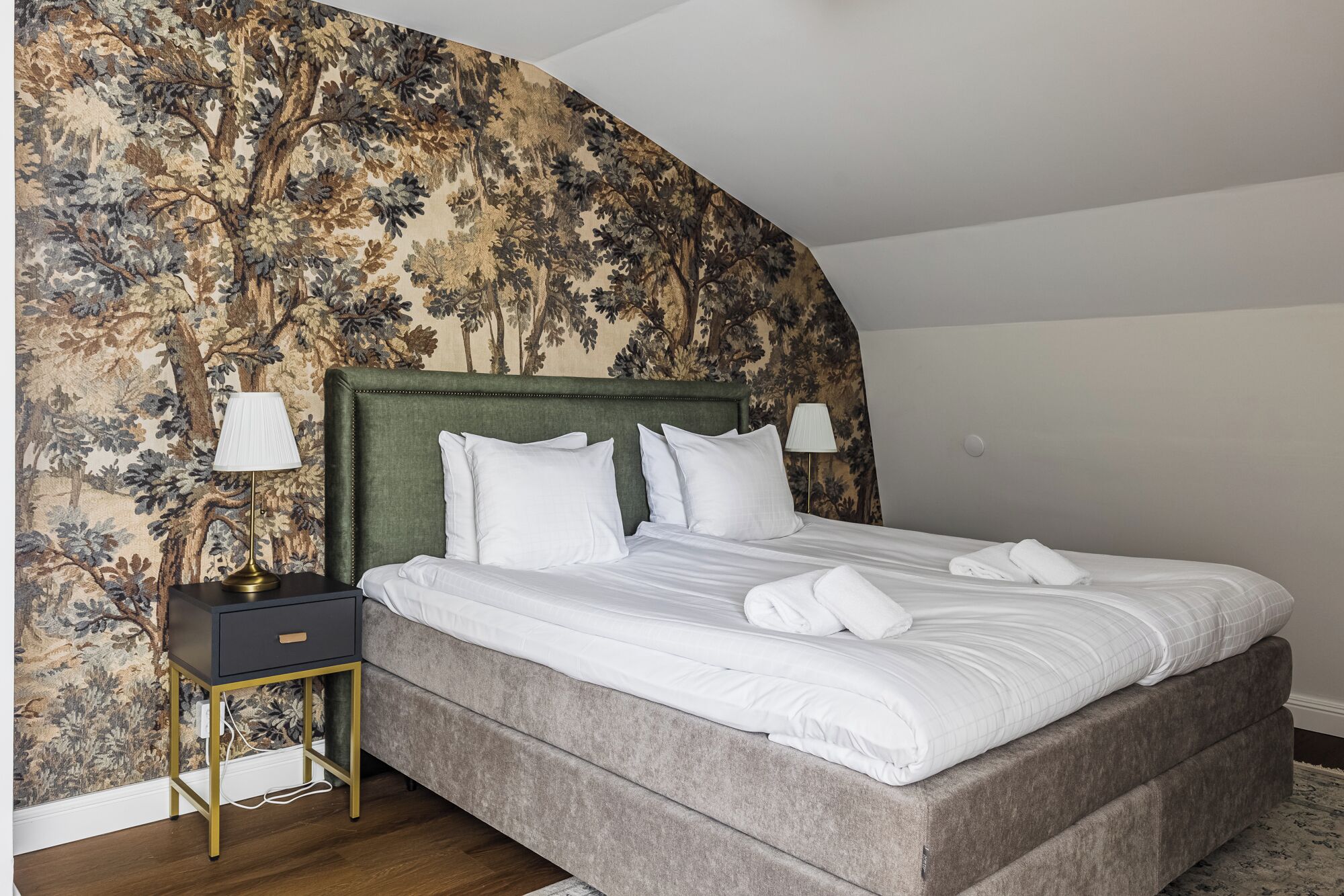 Quarto com uma cama grande, uma mesa de cabeceira com abajur e uma parede com padrão floral.