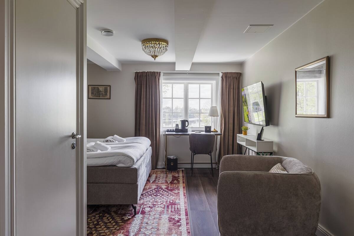 Habitación de hotel con cama, escritorio, silla, televisor y un amplio ventanal.