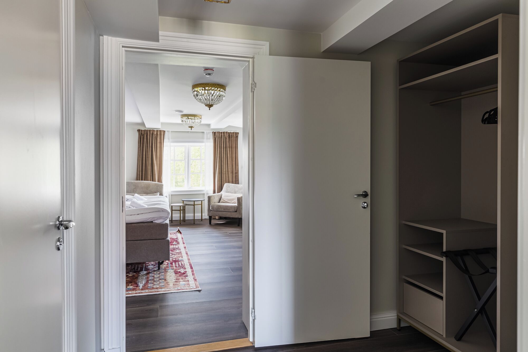 A hotel room with a bed, a chair, a small table, a wardrobe, and a chandelier.