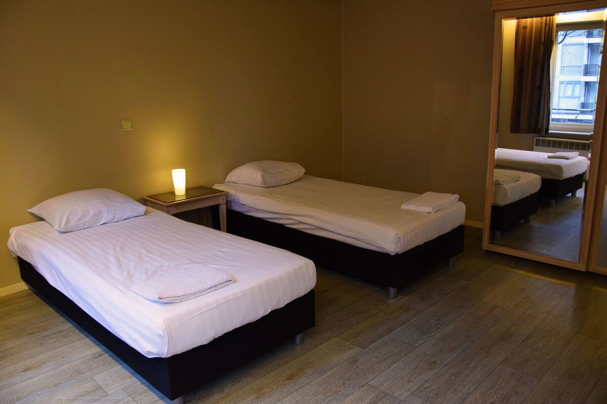 A hotel room with two single beds, a nightstand with a lamp, a large mirror, and wooden flooring.