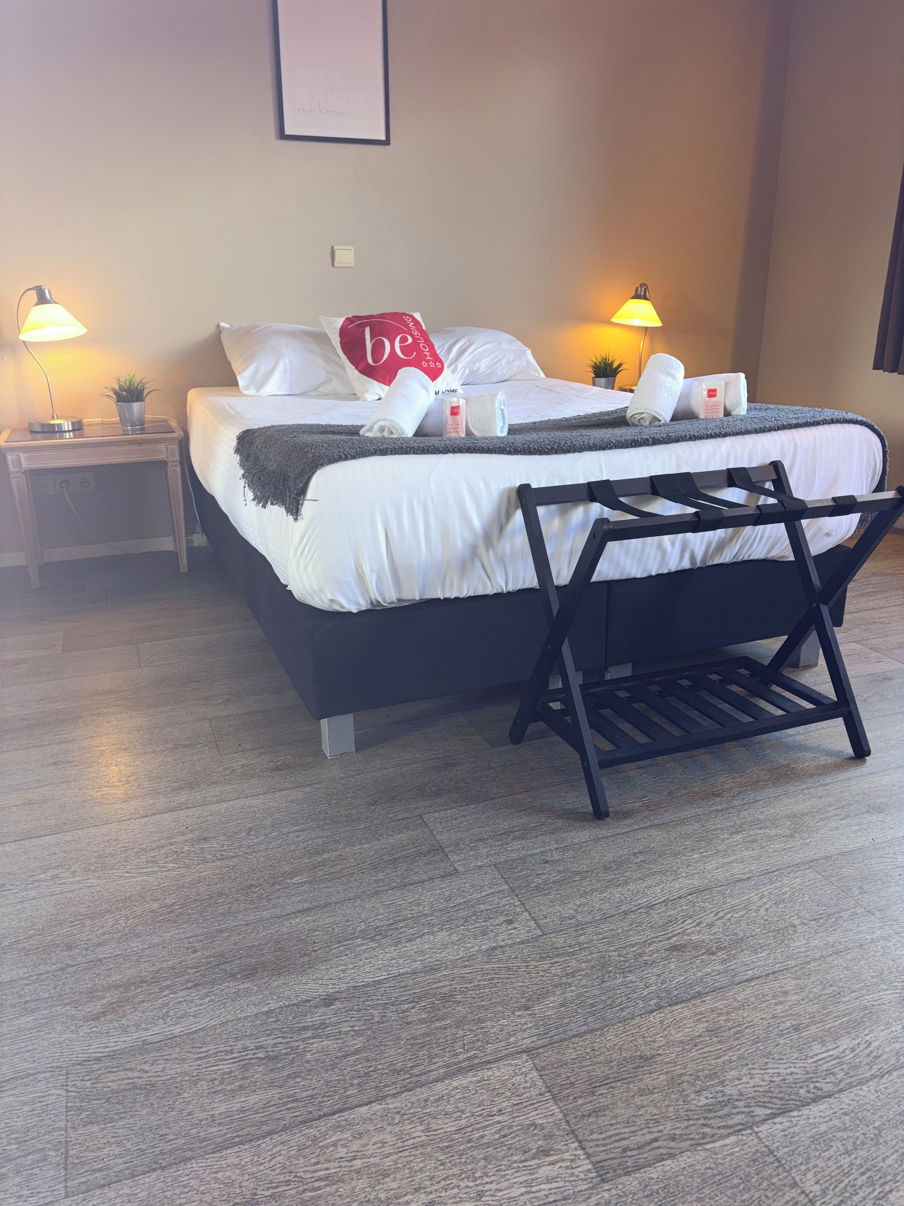 Een net opgemaakt bed met een grijze deken, een kussen met de tekst 'be' erop, en een nachtkastje met een lamp en een kleine plant.