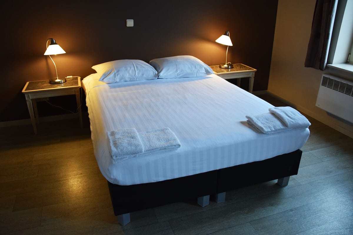 A neatly made bed with pillows and two folded towels, two bedside lamps, and a window with curtains.