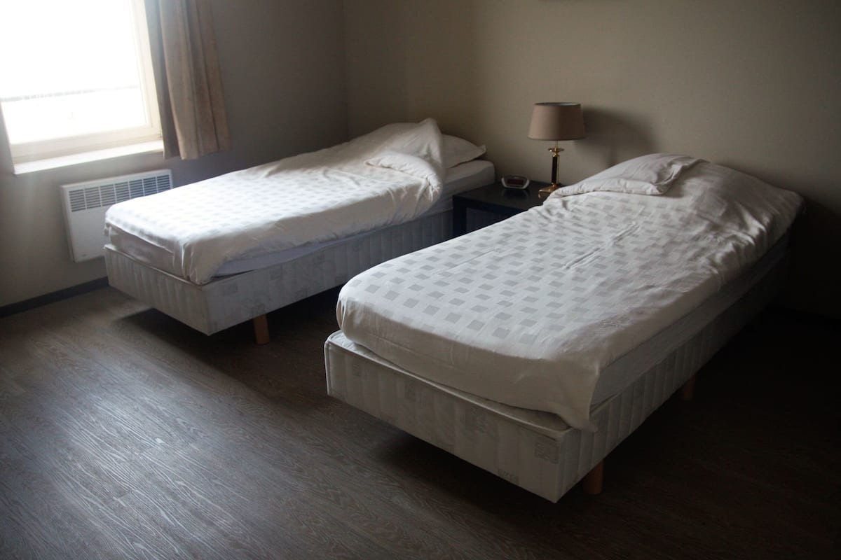 A room with two single beds, a bedside table with a lamp, and a window with curtains.