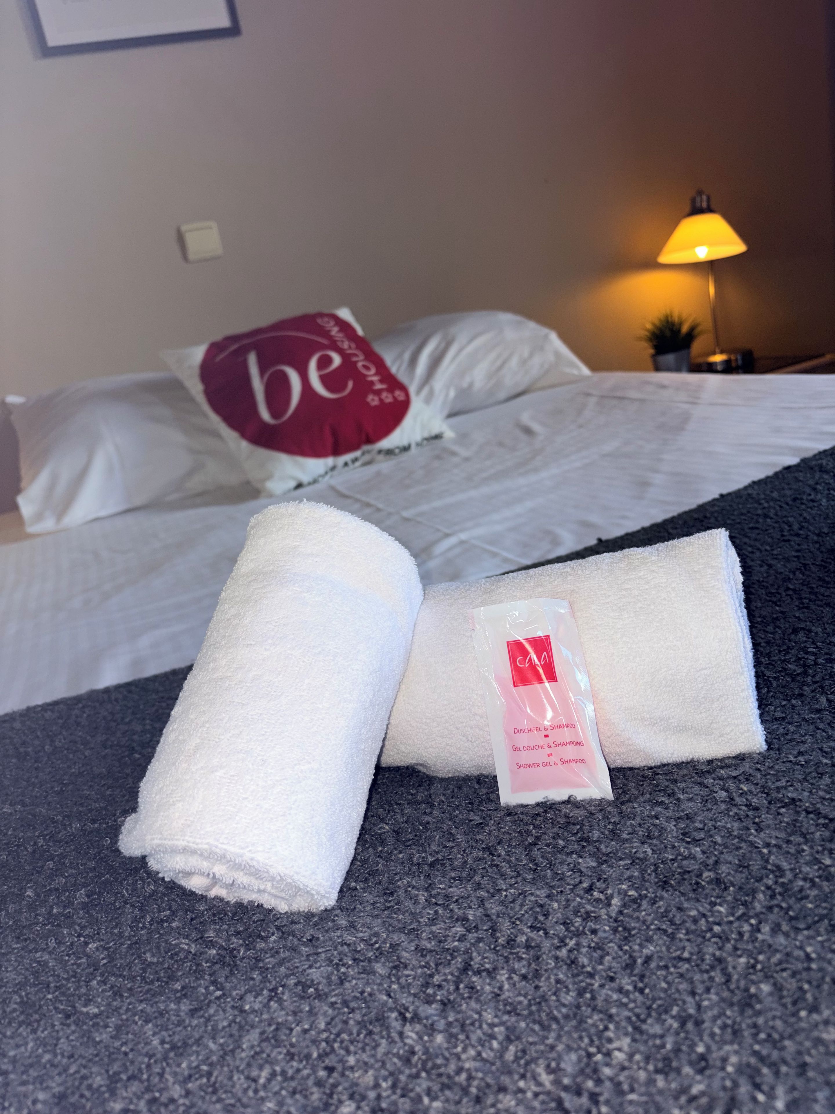 Una cama bien hecha con sábanas blancas y una almohada con la palabra 'be'. Hay dos toallas con etiquetas y una lámpara al lado.