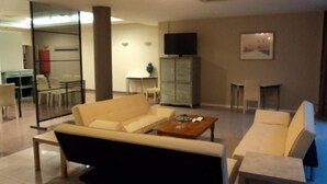 Una sala de estar moderna con sofá, mesa de centro y televisor. Se observa un comedor con sillas y una mesa al fondo.
