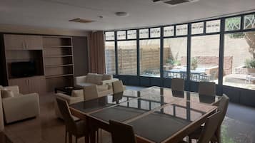 A modern living room with a glass-top dining table, sofas, a TV, and a view of the outside.