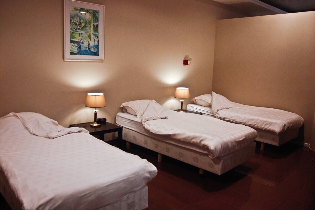 A hotel room with three single beds, each with a bedside table and lamp.