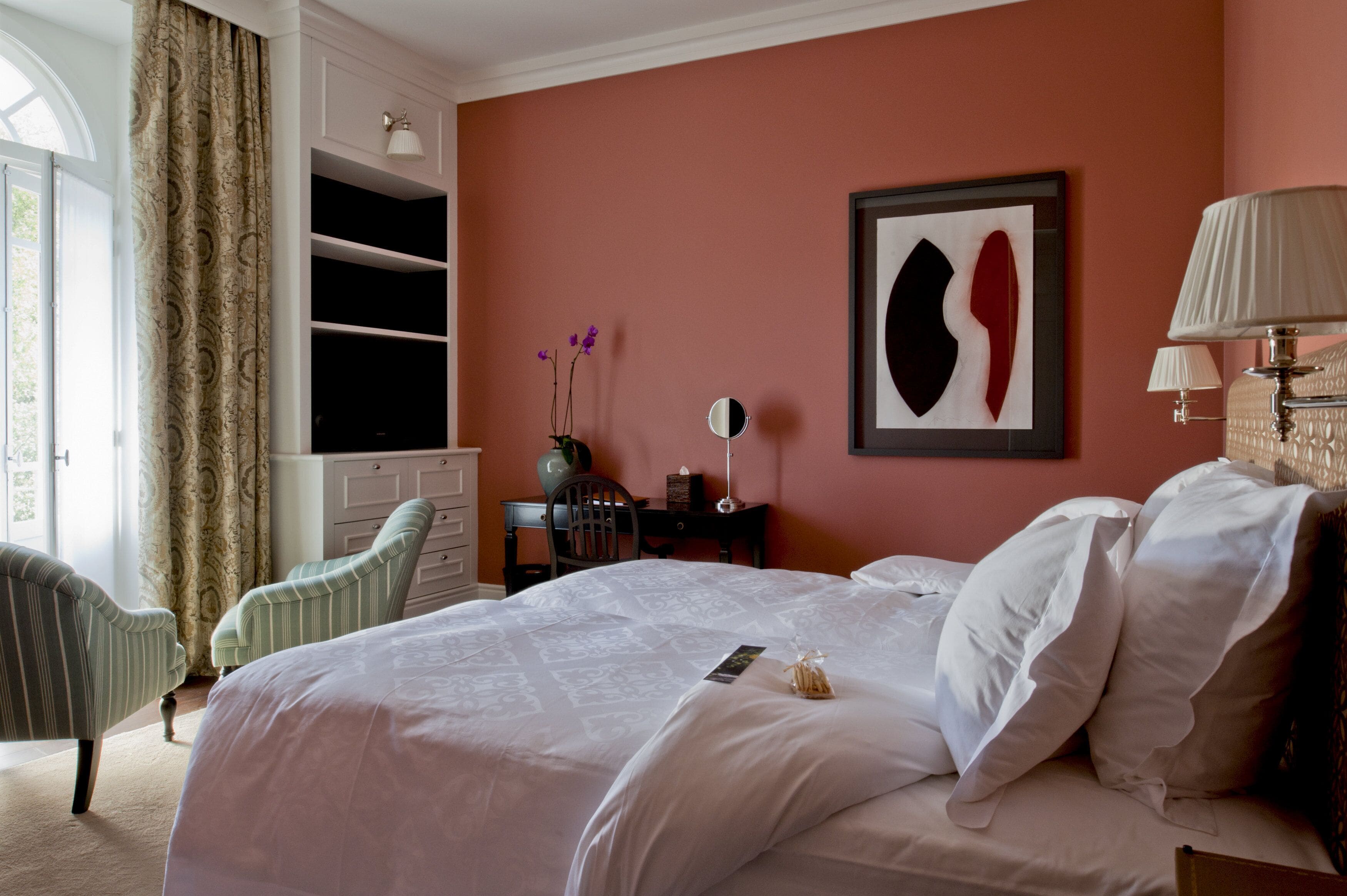 A hotel room with a large bed, a desk with a chair, a wardrobe, a mirror, and a window with curtains.