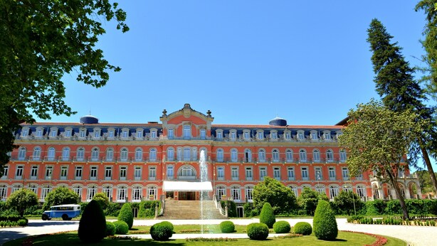Un grande edificio a più piani con una fontana centrale, circondato da giardini curati e alberi.