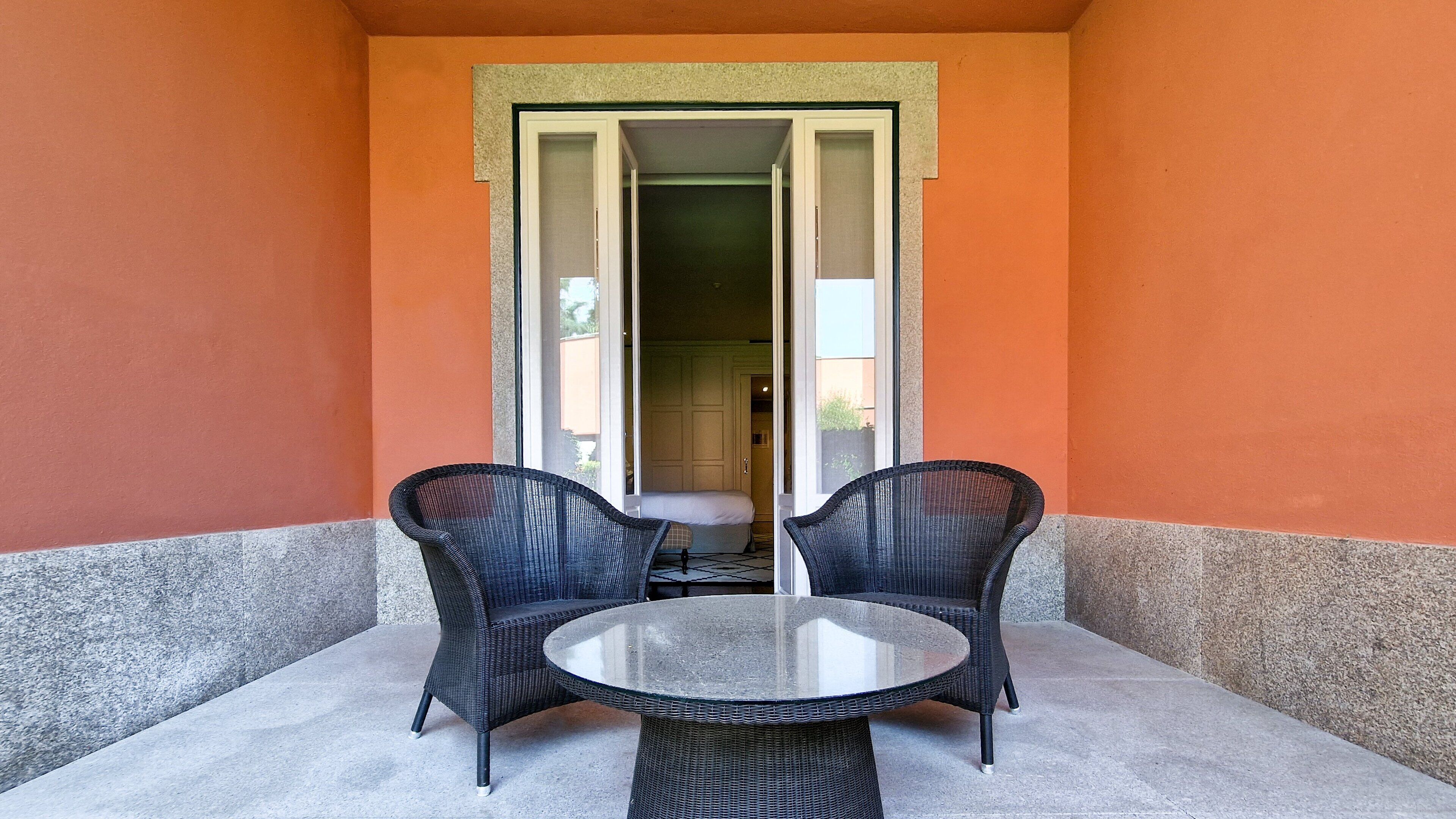 Un balcón con dos sillas de mimbre y una mesa de vidrio, una puerta que da a una habitación interior y vistas al exterior.