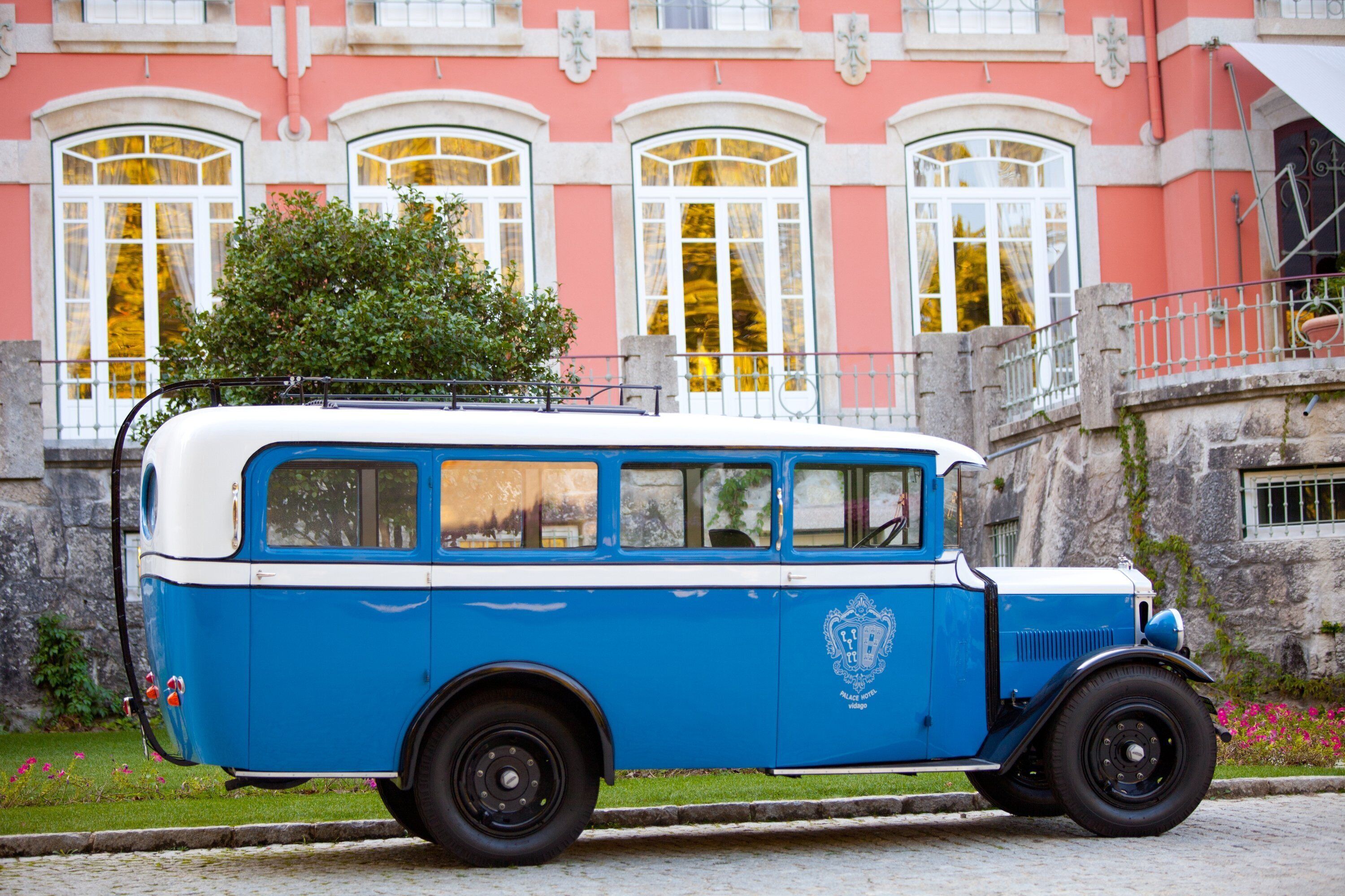 A vintage blue and white bus parked in front of a pink building with arched windows and a stone facade.