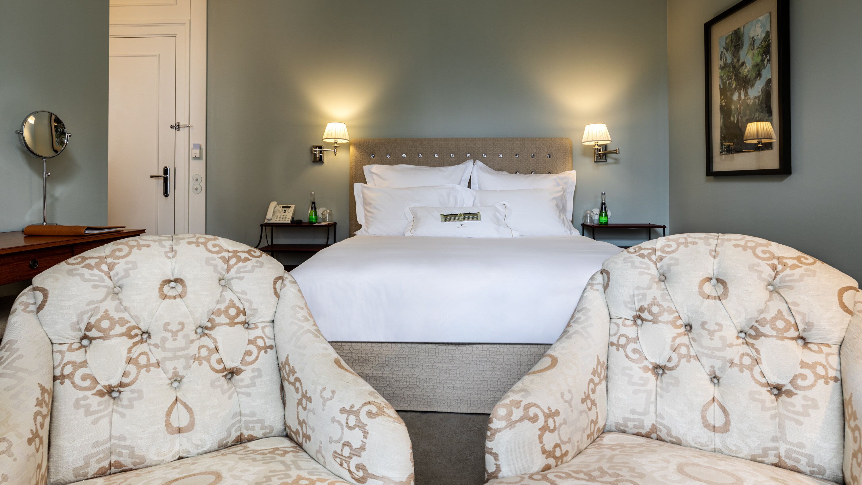 A hotel room with a large bed, two bedside tables, a mirror, and two upholstered chairs.