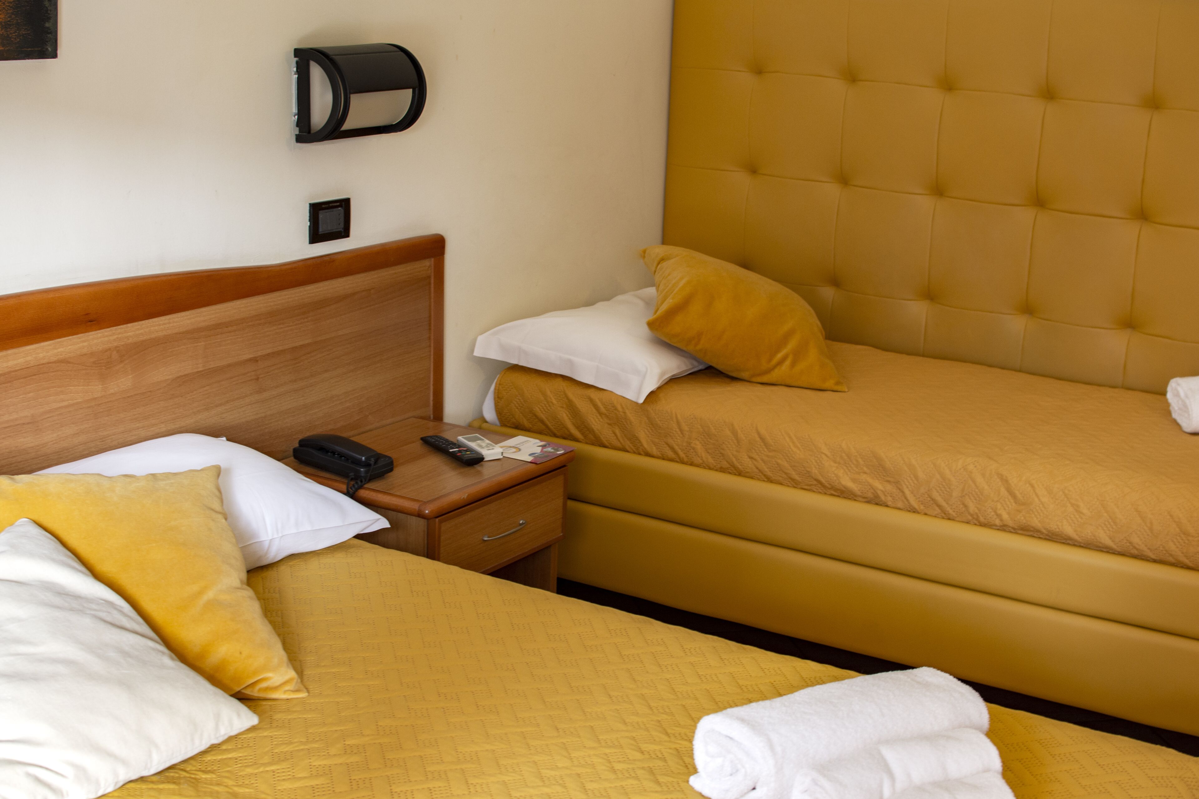 A hotel room with two beds, each with yellow bedding and white pillows. There is a wooden headboard, a nightstand with a phone and remote control, and a wall-mounted lamp.