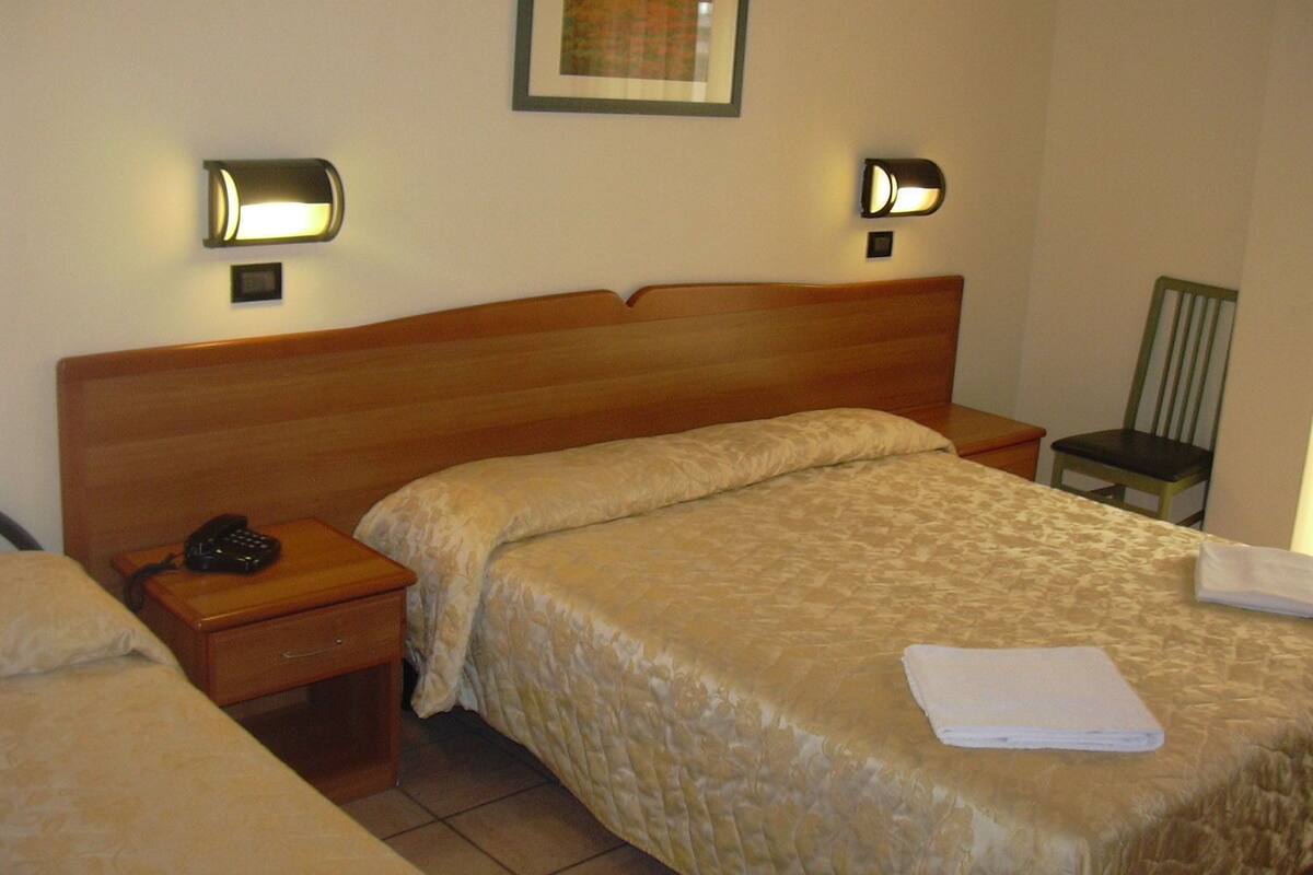 A hotel room with a wooden headboard, two bedside tables, a bed with a beige quilt, a chair, and two wall-mounted lamps.
