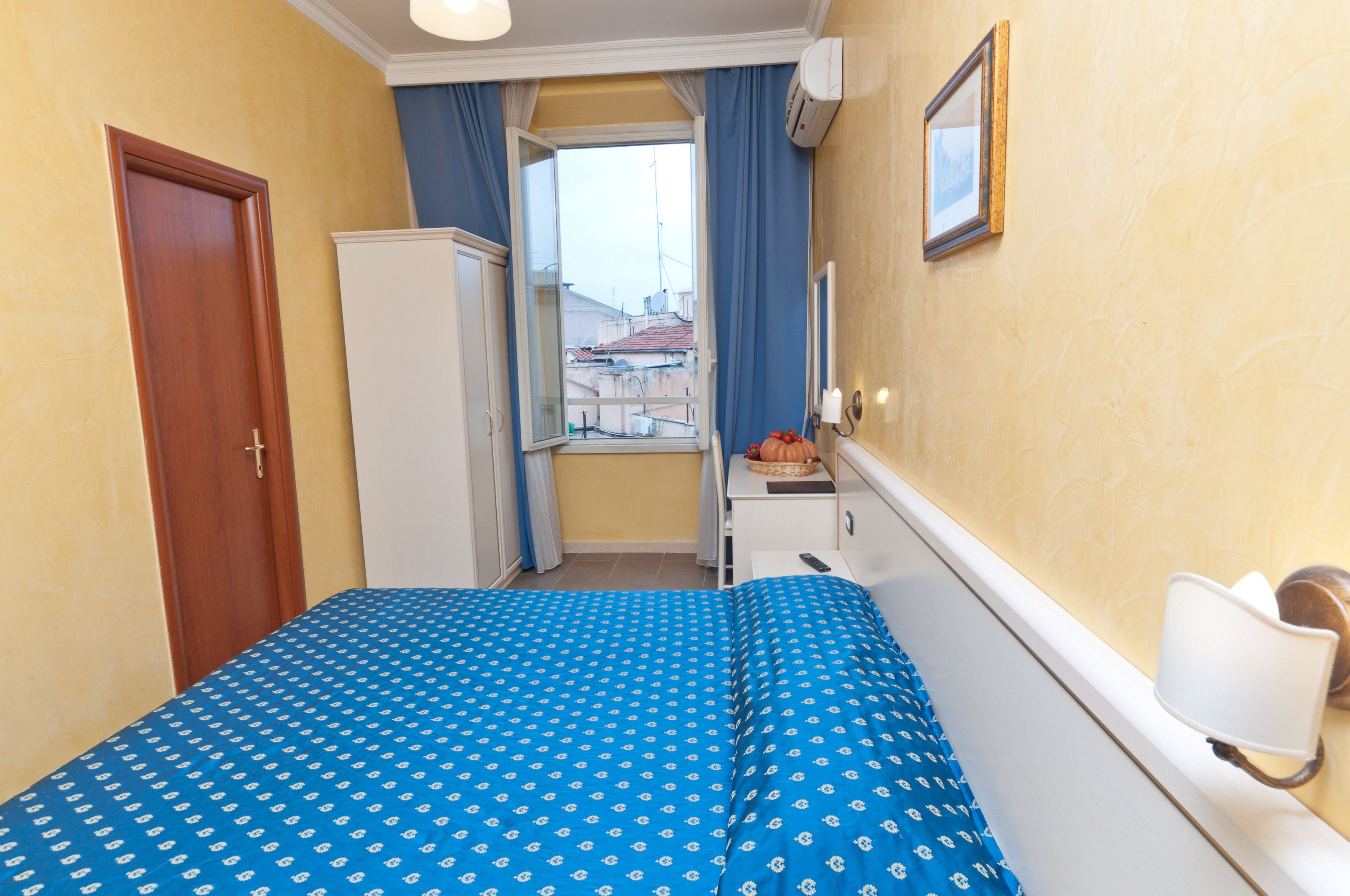 A hotel room with a bed, a wardrobe, a desk, a window with curtains, and a wall-mounted light fixture.