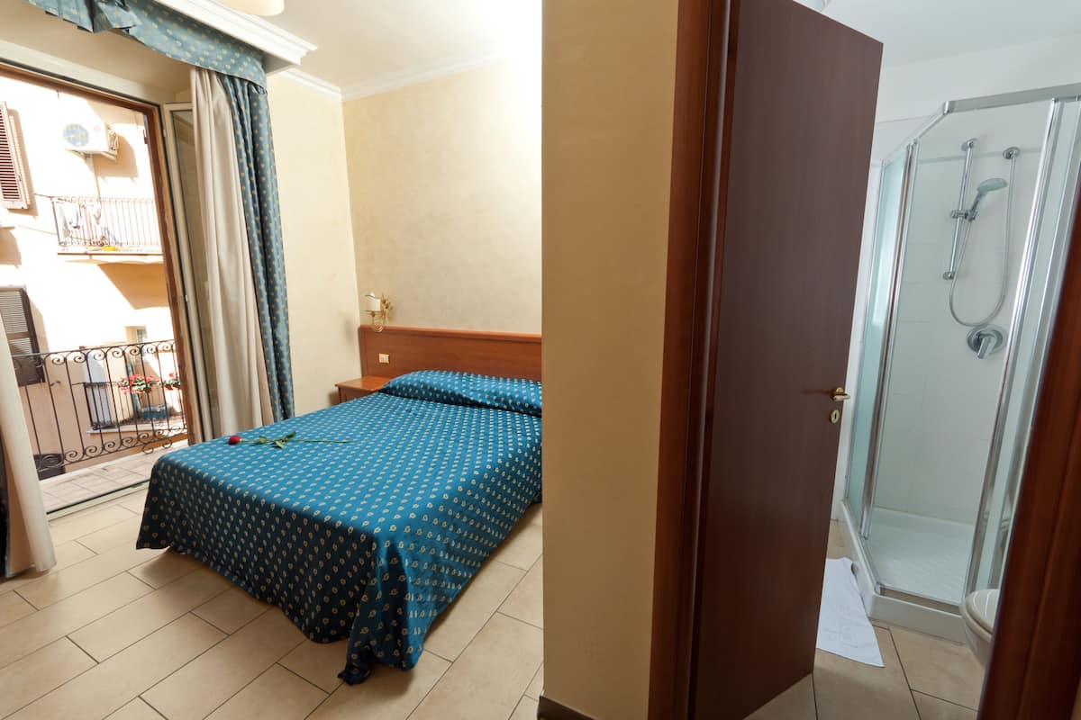 A room with a bed, a door leading to a shower, and a balcony with a railing.