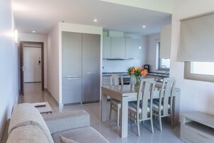 A modern living room with a sofa and a dining table with chairs. There is a kitchen area visible in the background with cabinets and a window.