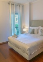 A neatly made bed with white linens and pillows, a bedside lamp, and a view of greenery through a window with curtains.