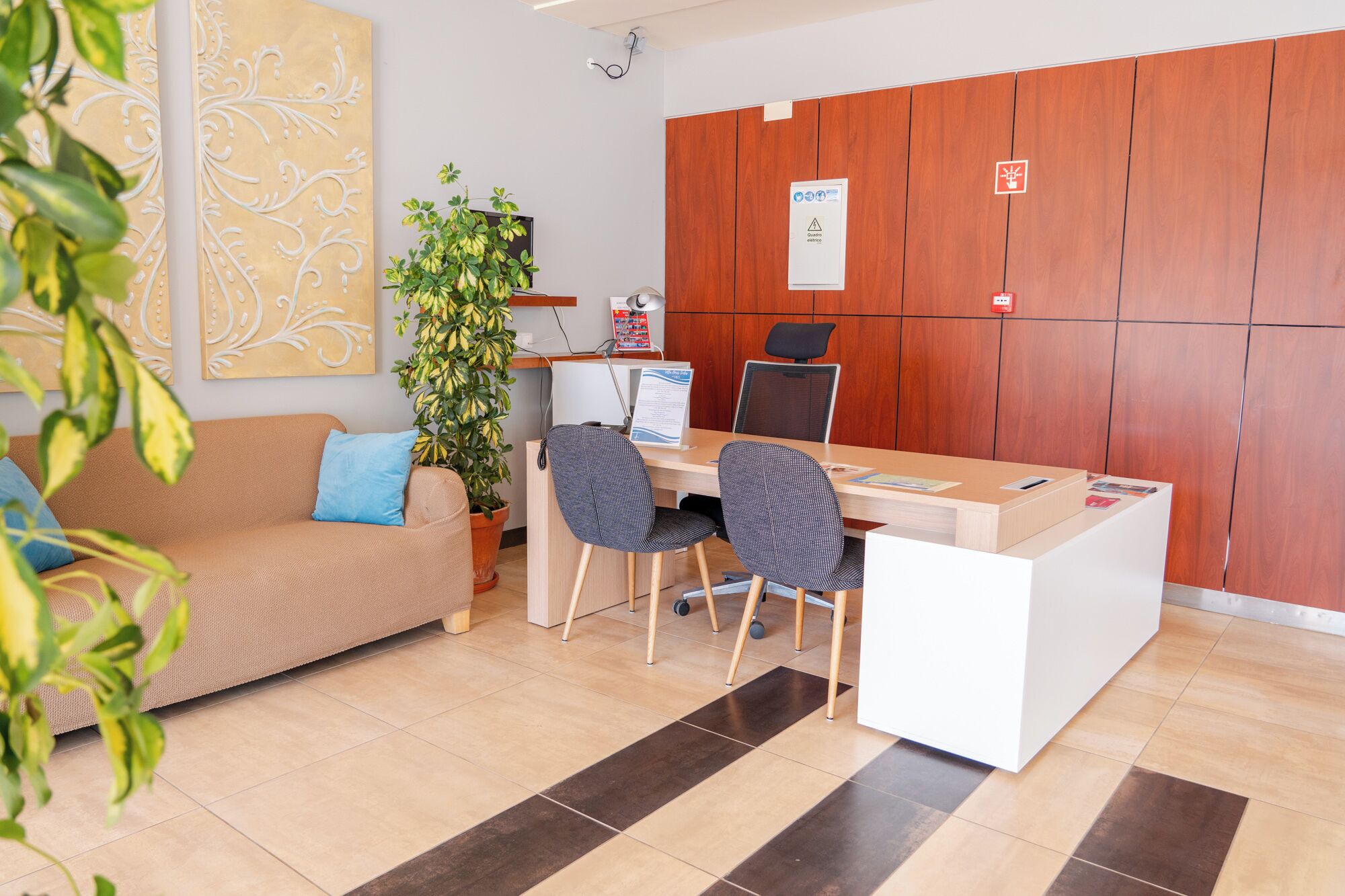 Un área de recepción con un panel de madera, un mostrador de recepción, dos sillas, un sofá con un cojín azul y una planta en maceta.