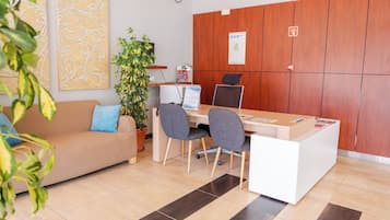 Un área de recepción con un panel de madera, un mostrador de recepción, dos sillas, un sofá con un cojín azul y una planta en maceta.