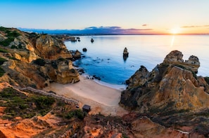 Un paisaje costero con acantilados rocosos, una playa de arena y un camino de madera que conduce a una pequeña construcción.