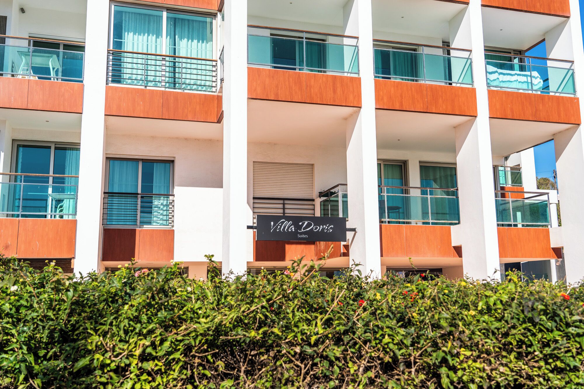 A modern hotel with multiple balconies, a sign for 'Villa Doris Suites', and a well-maintained hedge in the foreground.