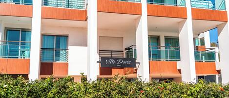 A modern hotel with multiple balconies, a sign for 'Villa Doris Suites', and a well-maintained hedge in the foreground.