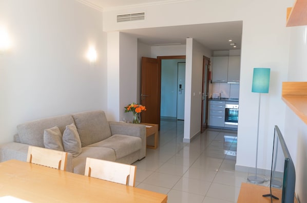 Una sala moderna con sofá, mesa de comedor y sillas. Un florero con flores está sobre la mesita de noche. Se ve la cocina al fondo.