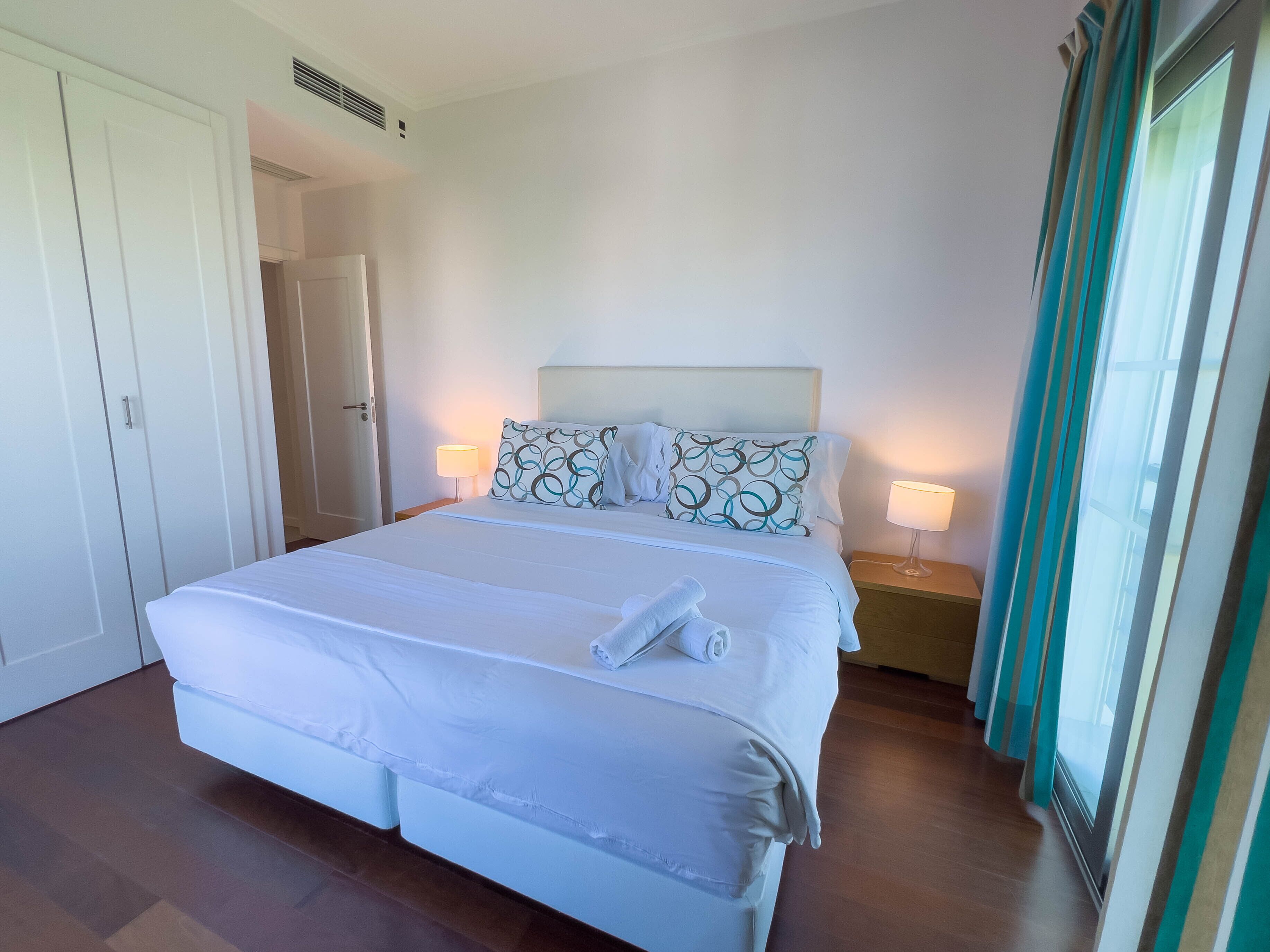 A neatly made bed with white linens and two pillows, a bedside table with a lamp, and a view of a window with curtains.
