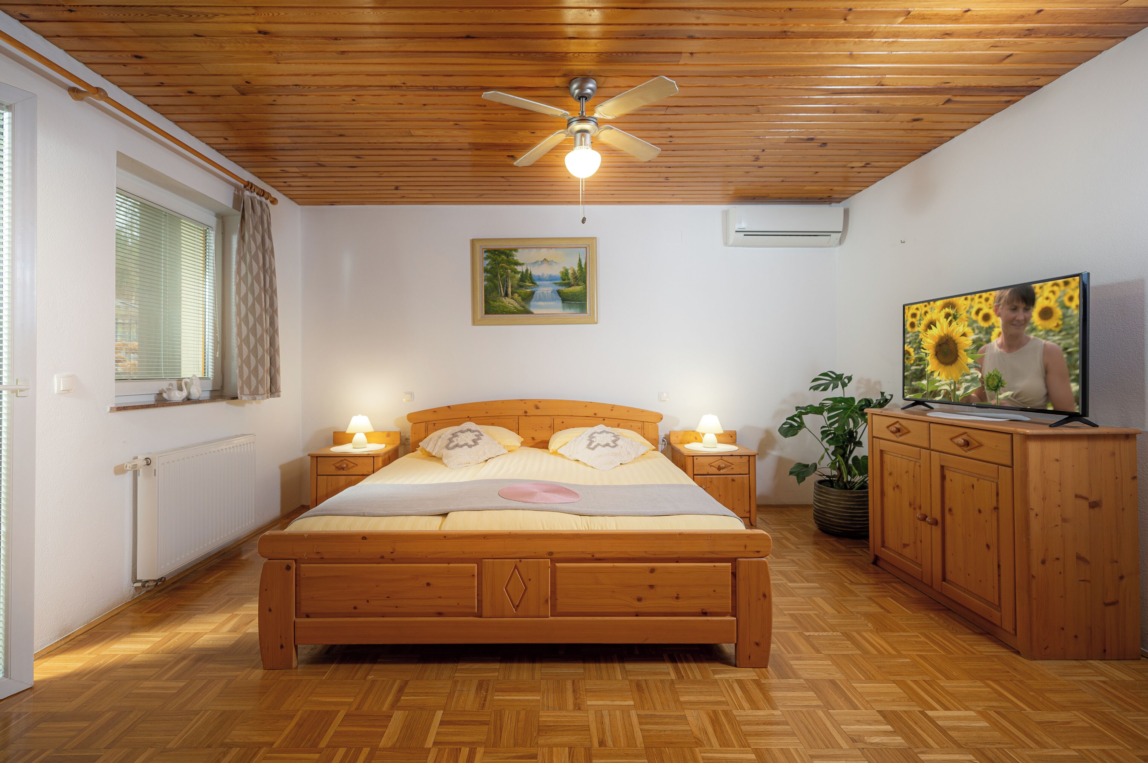 A bedroom with a wooden bed, bedside lamps, a ceiling fan, a painting, a TV, and a potted plant.