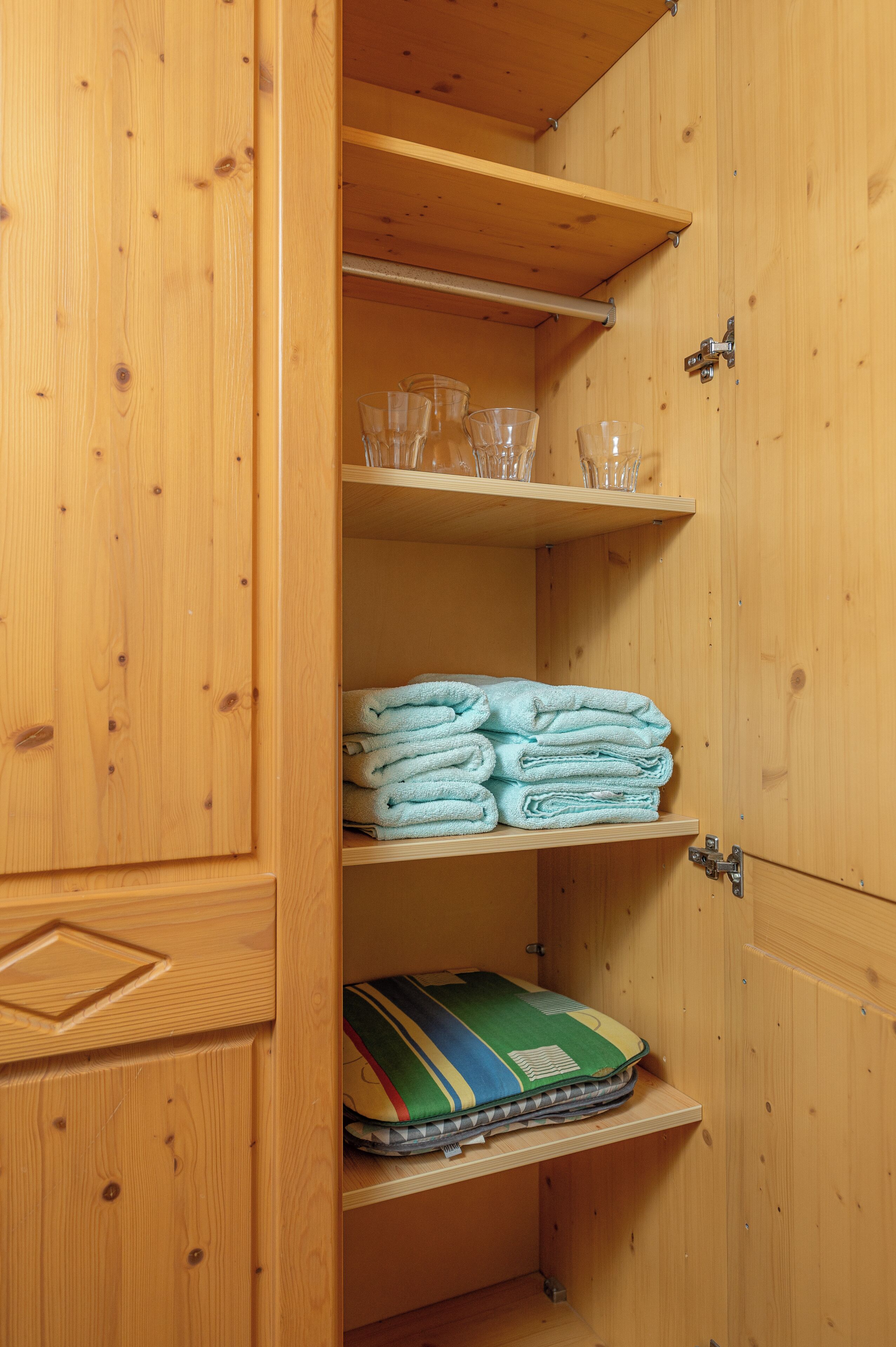 A wooden wardrobe with shelves holding folded towels, glasses, and a pillow.