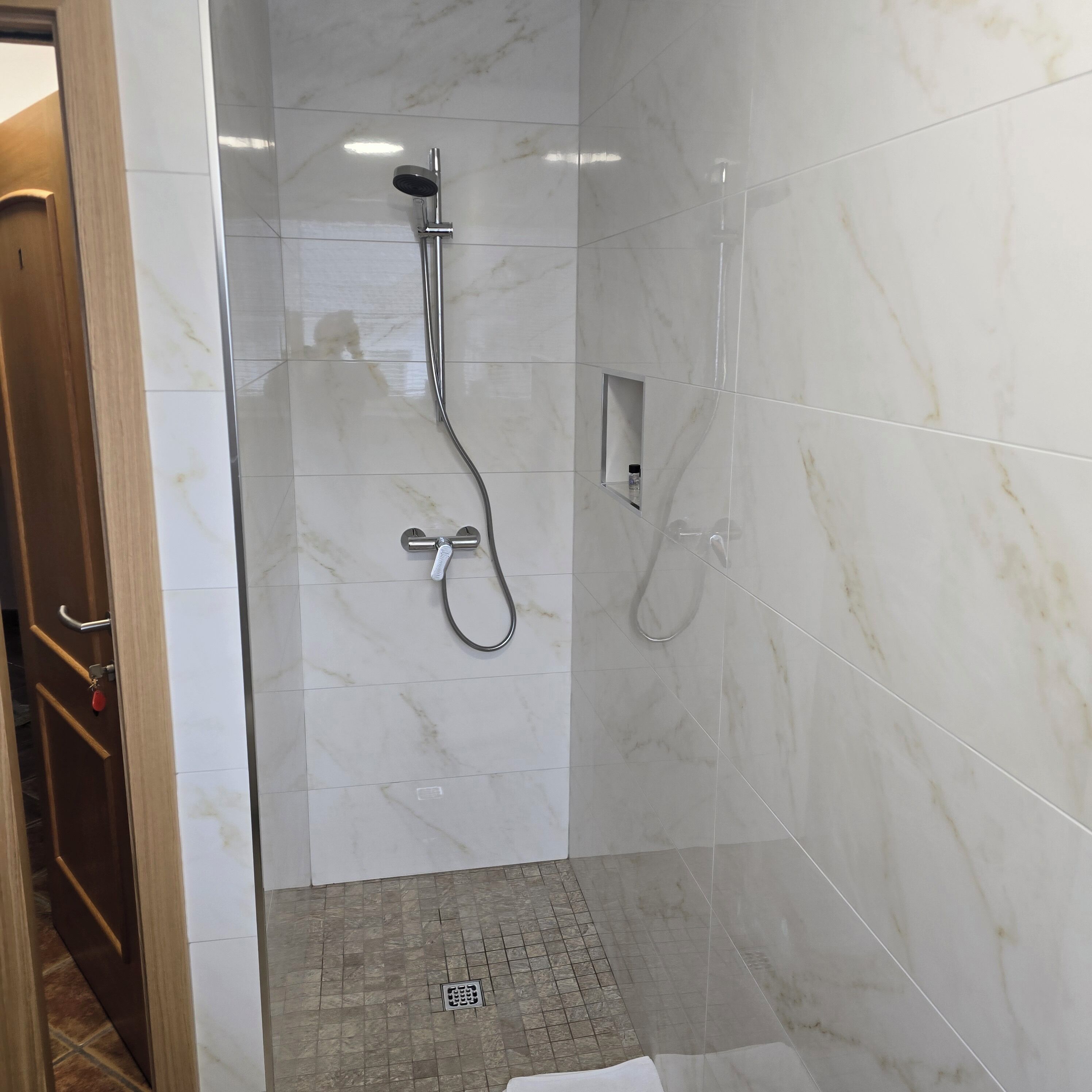 A shower with a handheld showerhead and a wall-mounted faucet, a small mirror, and a tiled floor with a drain.