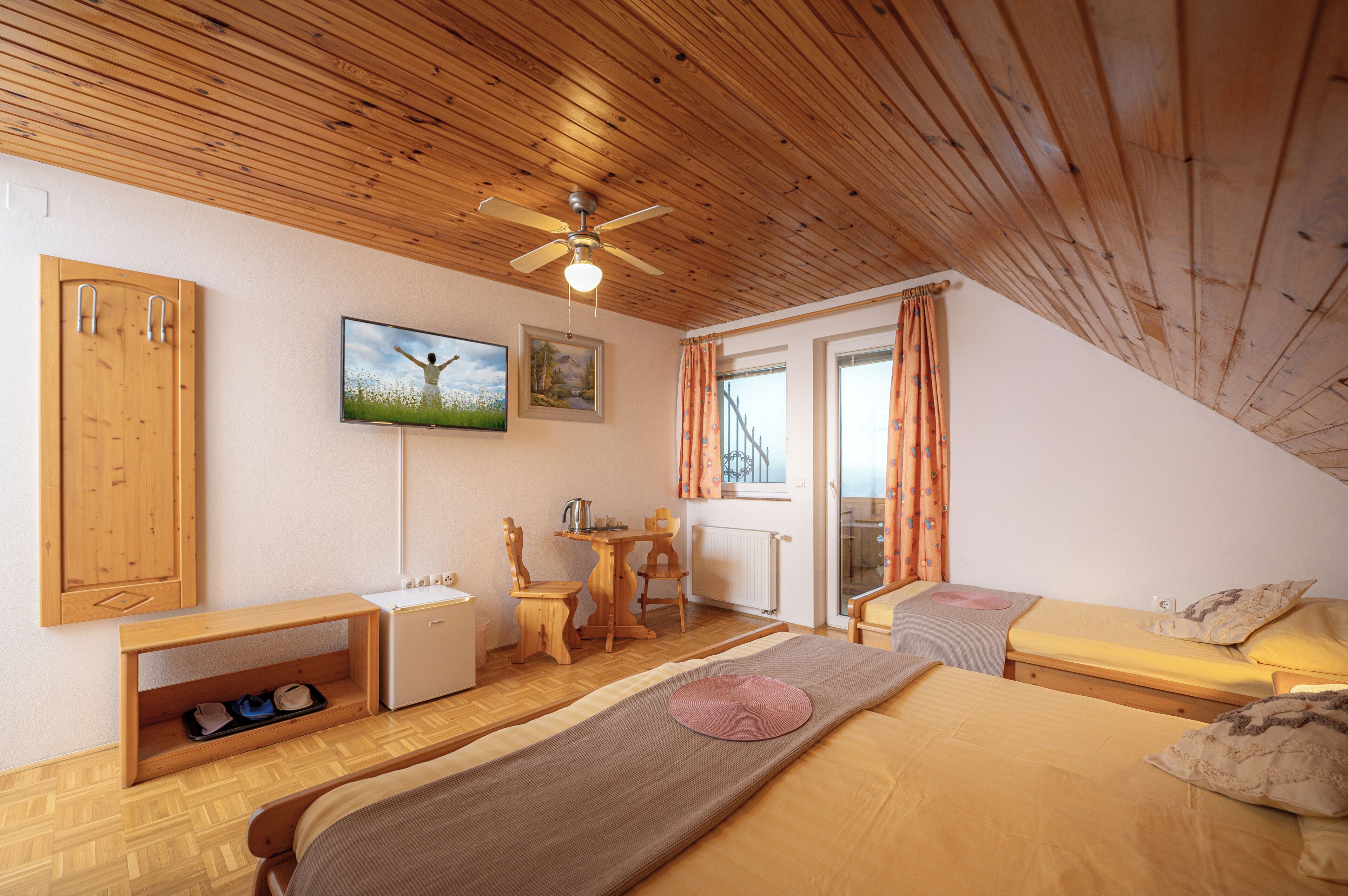 A room with a wooden ceiling, a bed, a desk, a chair, a radiator, and a window with curtains.