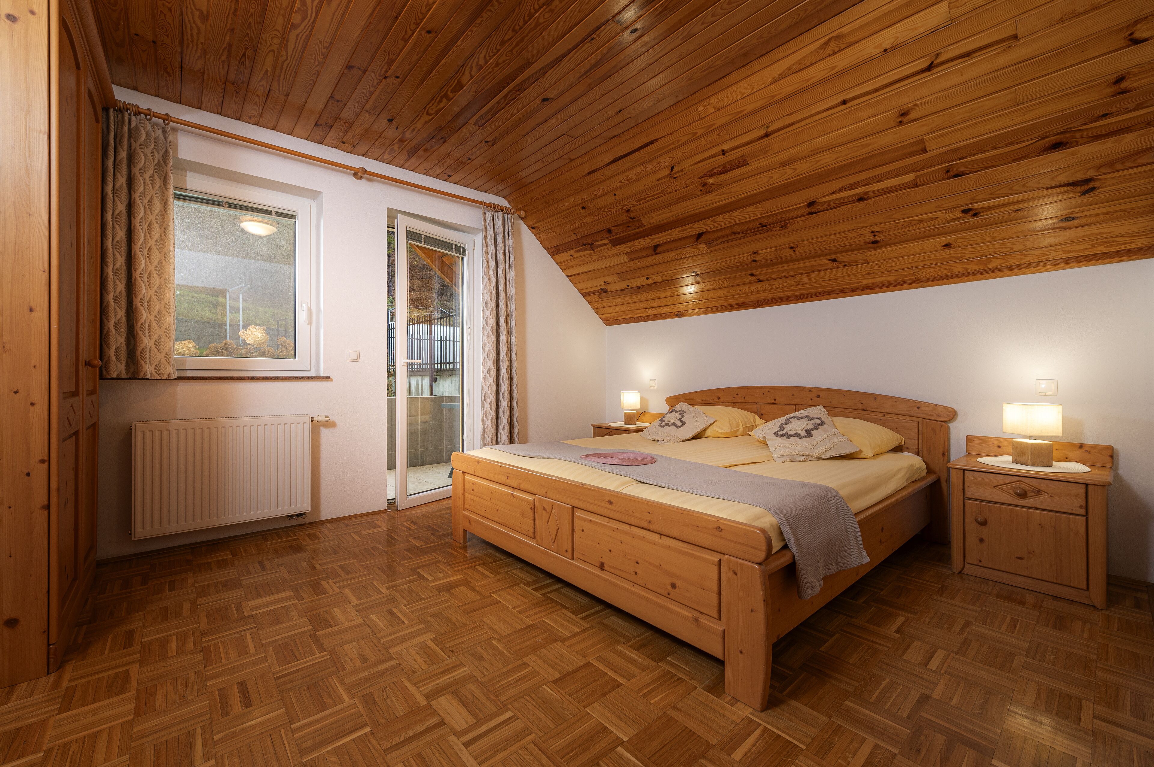 A bedroom with a wooden bed, bedside tables with lamps, a radiator, and a sliding glass door leading to a balcony.