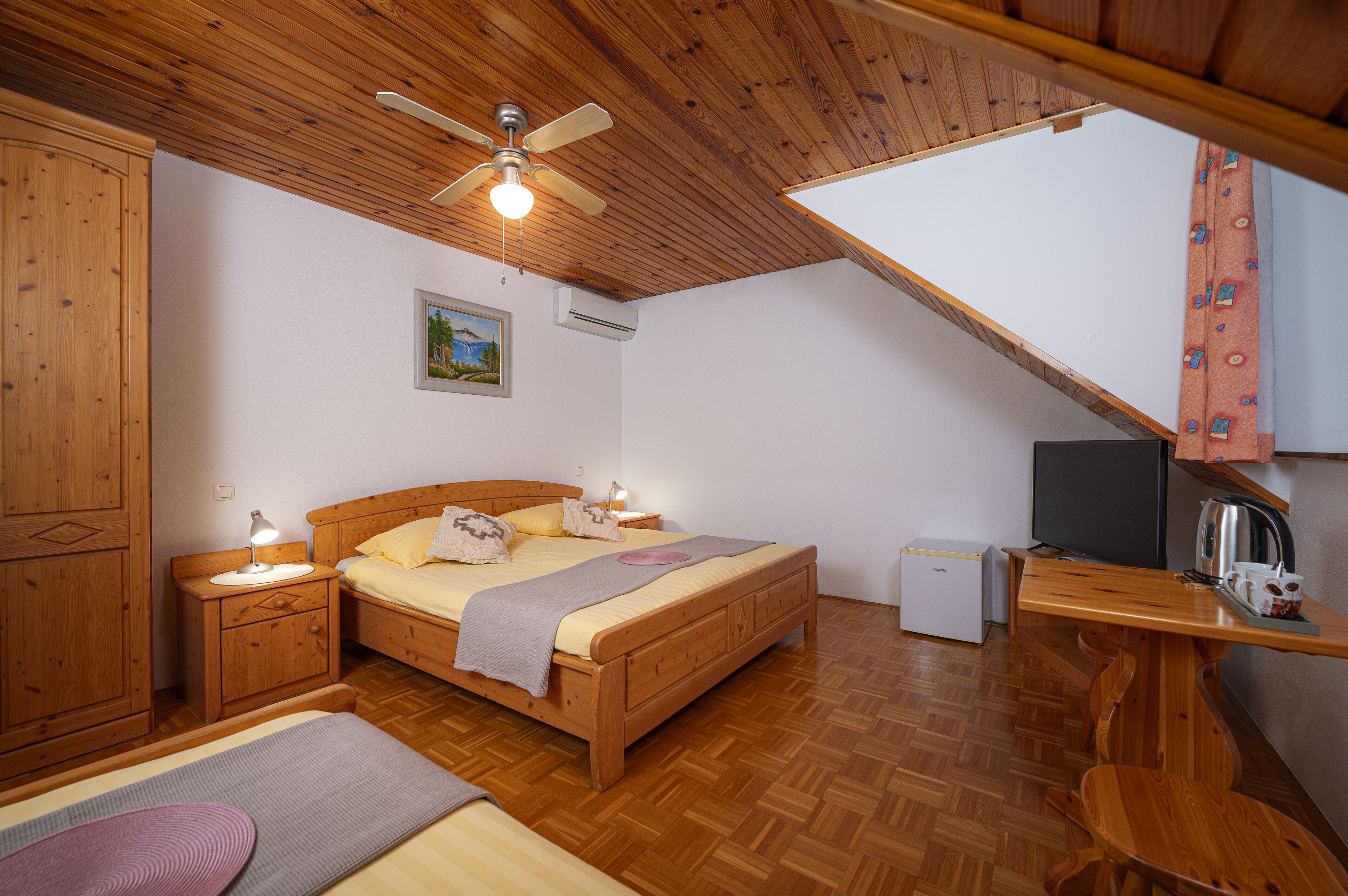 Ein gemütliches Schlafzimmer mit Holzbalkendecke, einem Bett mit Nachttisch, einem Schreibtisch mit Stuhl und einer kleinen Küchenzeile.