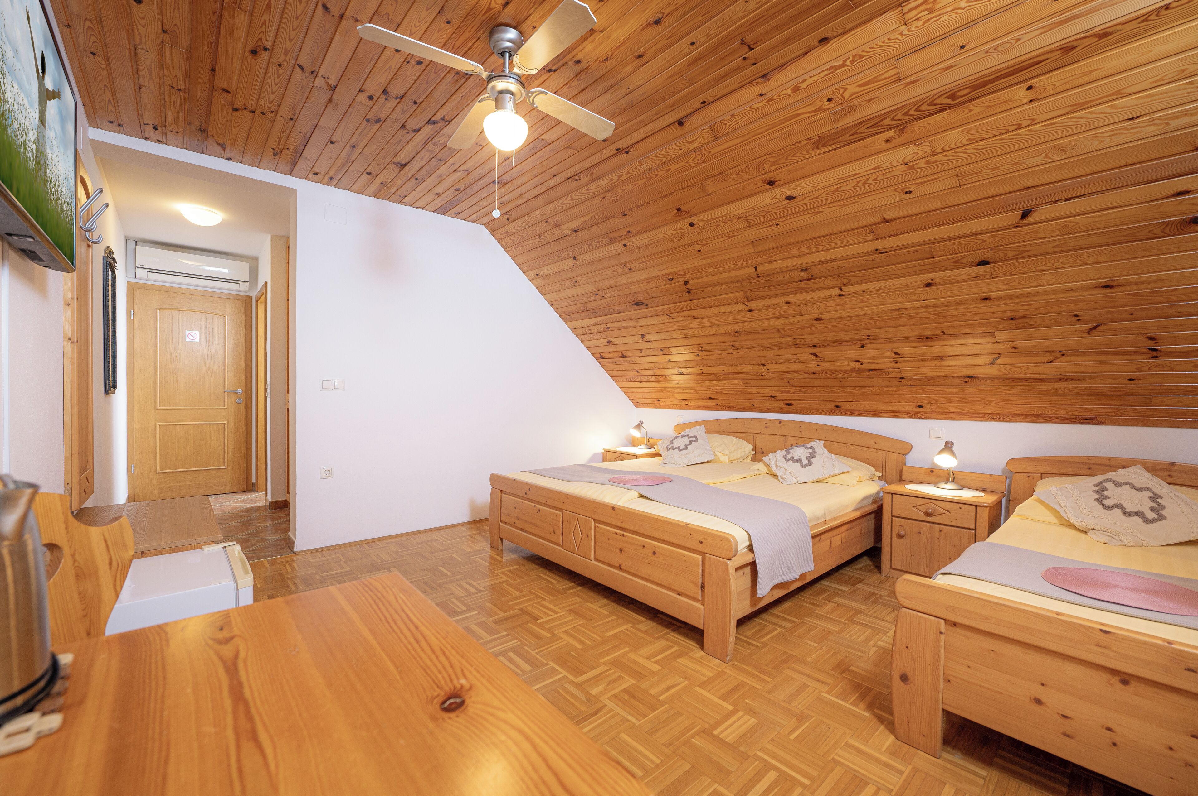 Un dormitorio con dos camas de madera, un ventilador de techo, una mesa de madera y una puerta que da a otra habitación.
