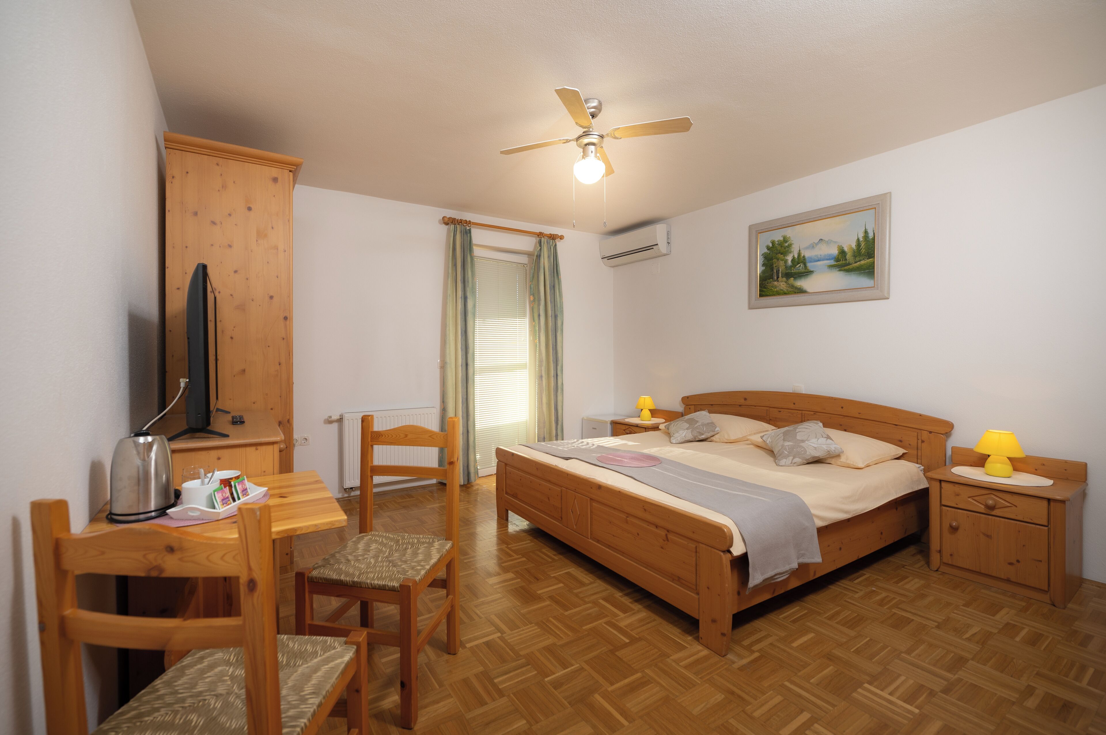 A bedroom with a bed, bedside tables, a desk, a chair, a wardrobe, a ceiling fan, and a window with curtains.