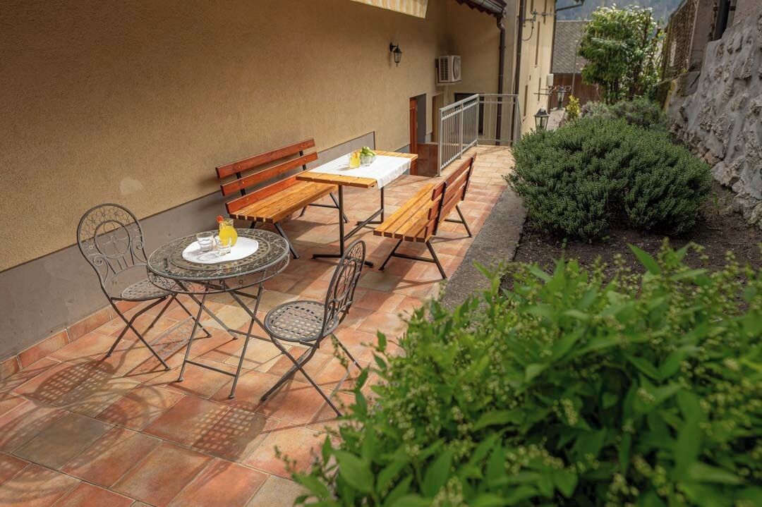 Un patio al aire libre con una mesa y bancos, rodeado de follaje y un muro de piedra.
