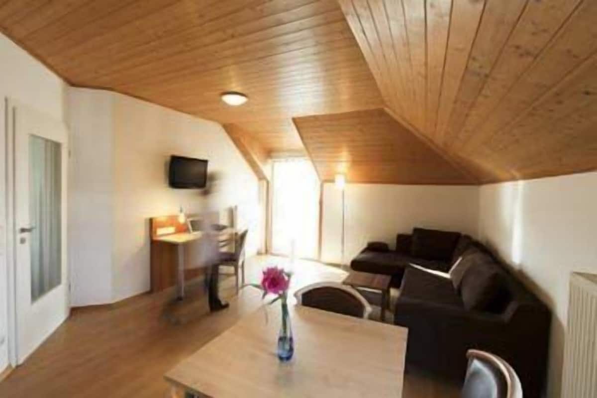 A cozy loft-style room with a wooden ceiling, a dining area, a sofa, and a TV mounted on the wall.