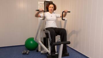 Uma pessoa usando uma máquina de leg press em uma academia, com uma bola de exercícios verde e halteres no chão.