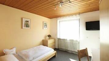 Quarto pequeno e simples, com uma cama, uma escrivaninha e uma cadeira. Há uma janela com vista para o exterior.