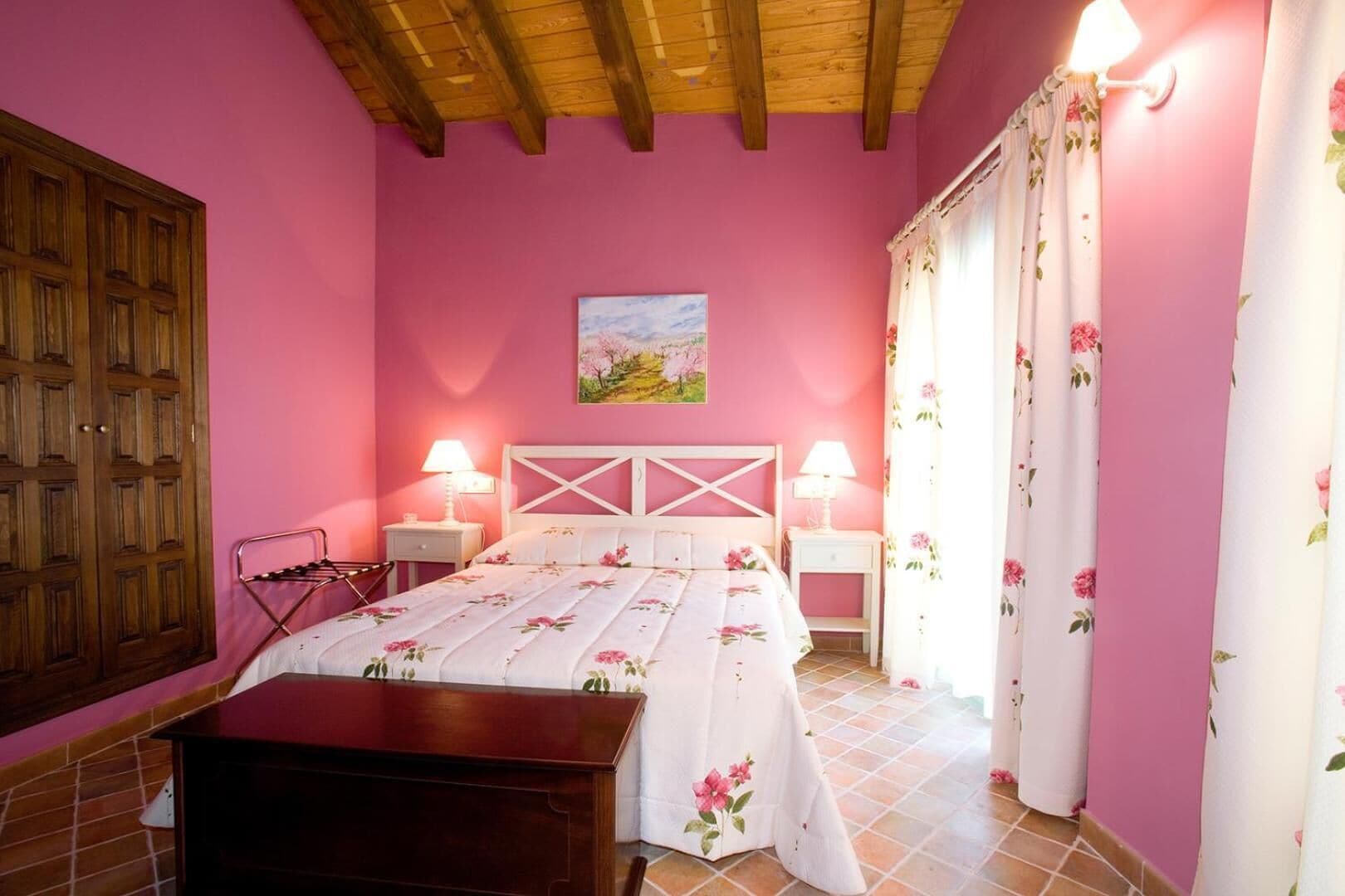 A bedroom with pink walls, a wooden ceiling, a bed with floral bedding, two bedside lamps, and a wooden wardrobe.