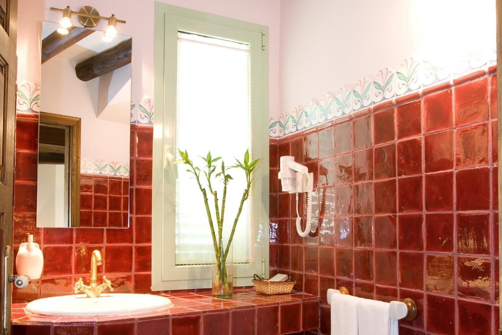 A bathroom with a red-tiled wall, a white sink with a gold faucet, a mirror, a window with blinds, and a hairdryer on the wall.