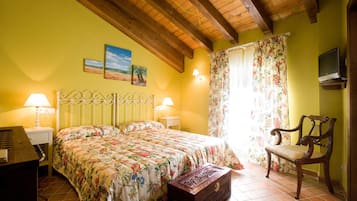 A bedroom with a bed, two bedside lamps, a wooden chair, a television, and a window with floral curtains.