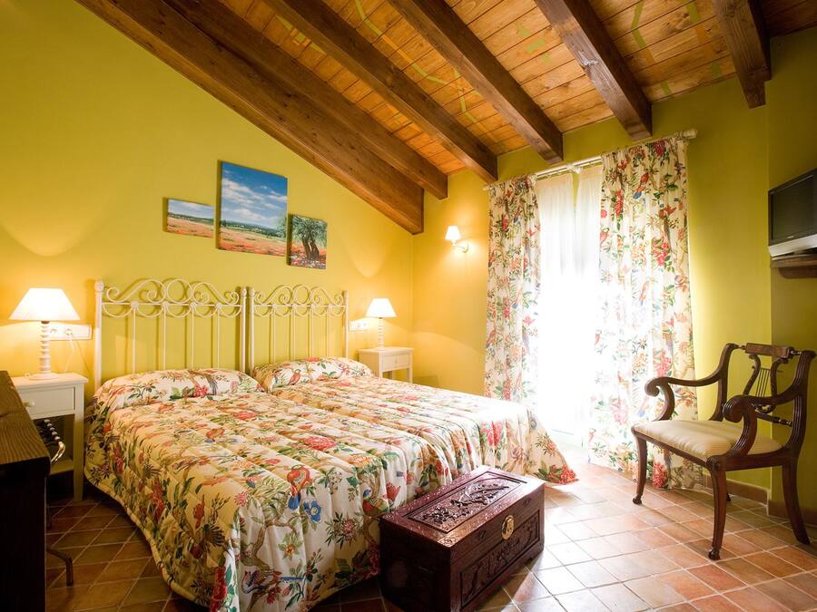 A bedroom with a bed, two bedside lamps, a wooden chair, a television, and a window with floral curtains.
