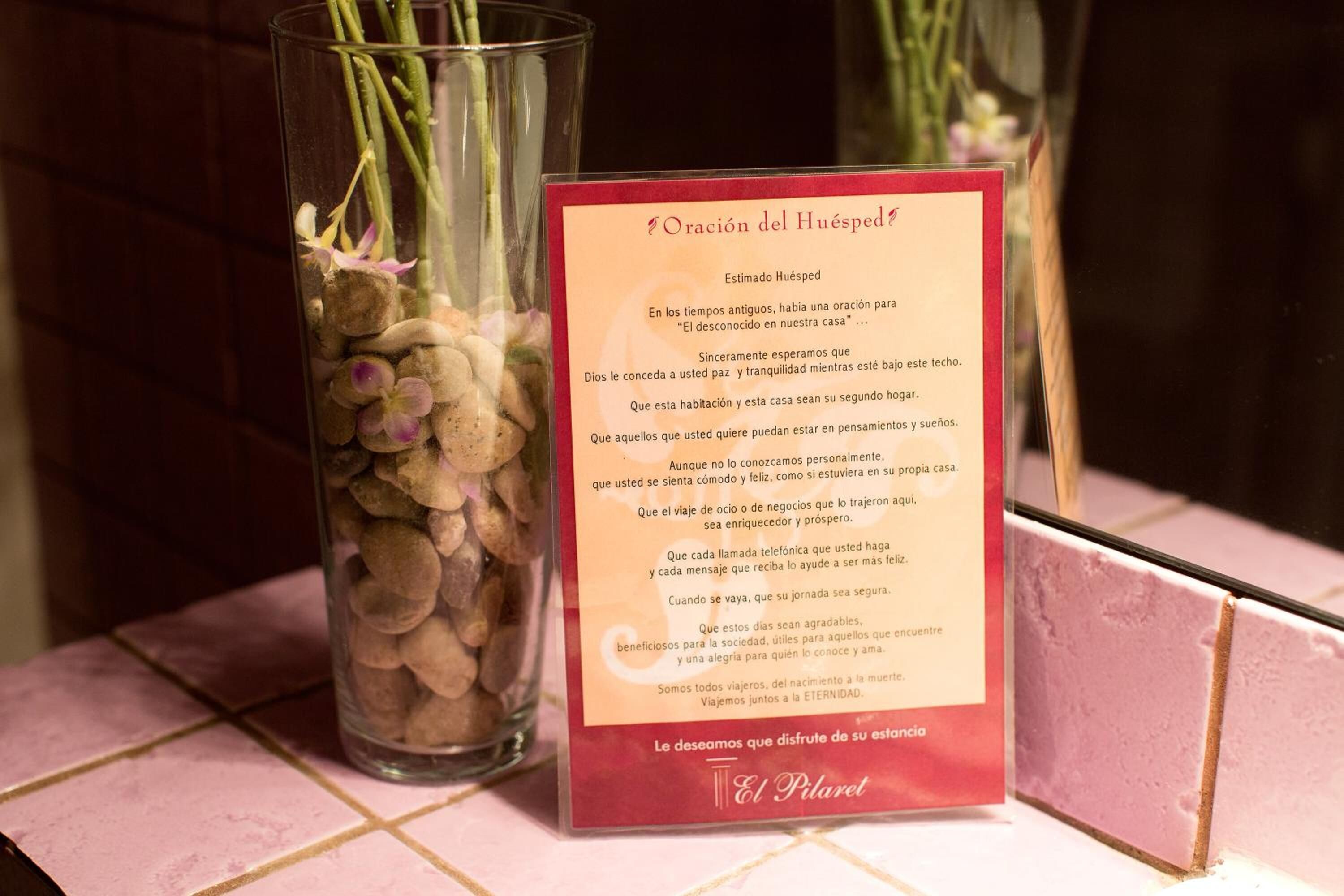 A prayer card titled "Oración del Huésped" next to a vase with flowers and stones.