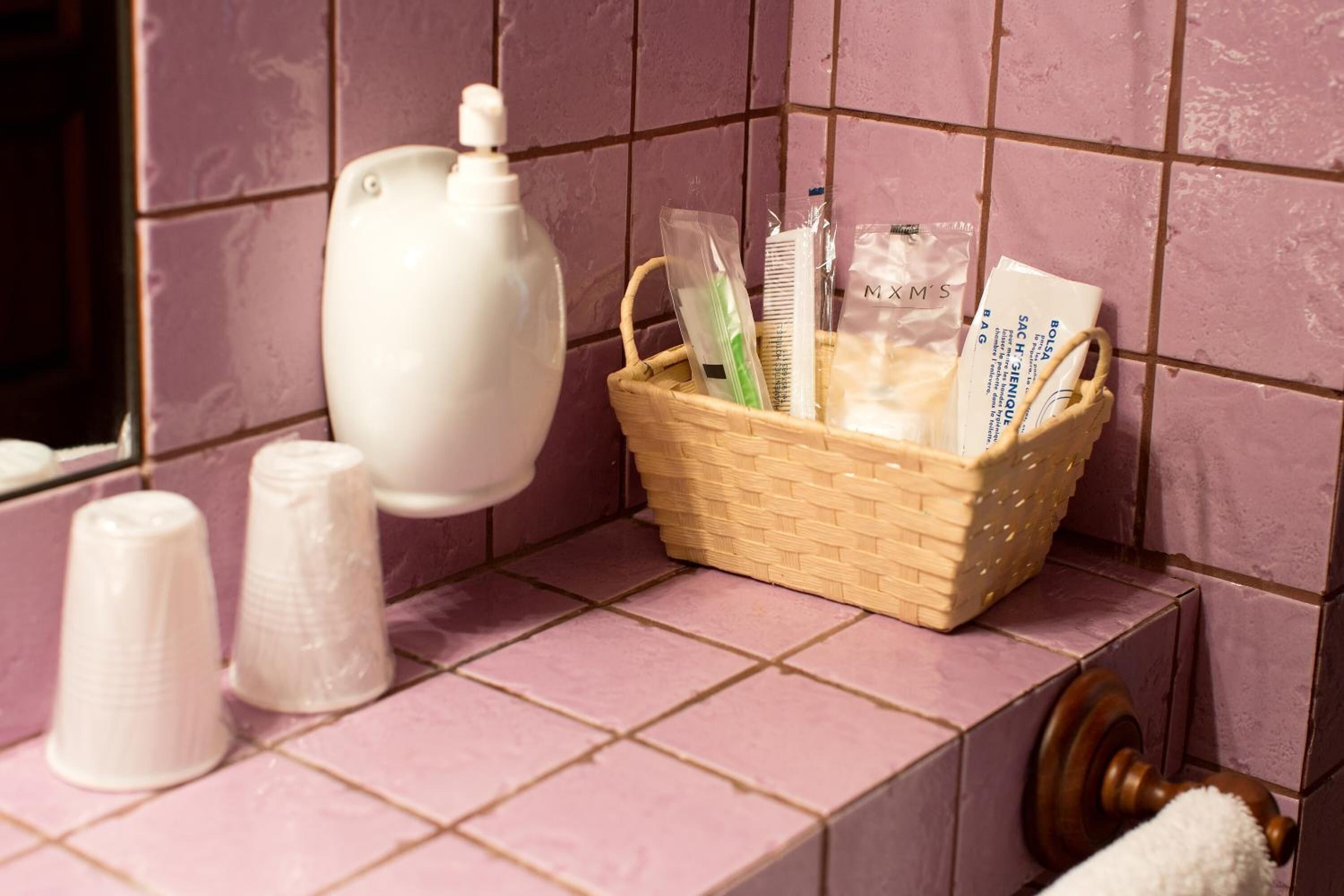 A bathroom with a pink tiled wall, a white soap dispenser, a wicker basket containing toiletries, and a towel rack.
