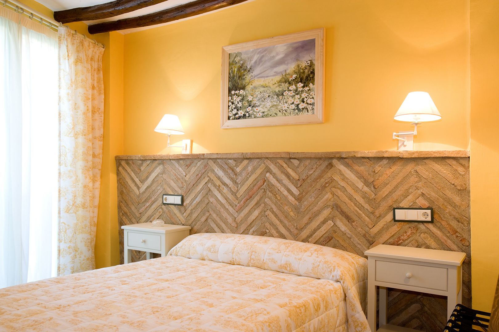 A bedroom with a herringbone-patterned headboard, two bedside tables, a bed with a floral bedspread, and a painting of a landscape on the wall.